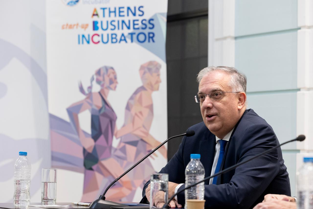 A man in a suit speaks at a conference, with a banner for the Athens Business Incubator in the background. Water bottles are on the table.