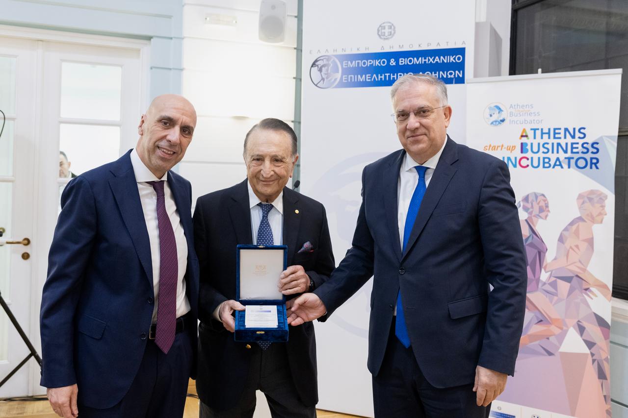 Three men are standing together, one holding a medal in a blue box. They are in a formal setting with banners in the background.