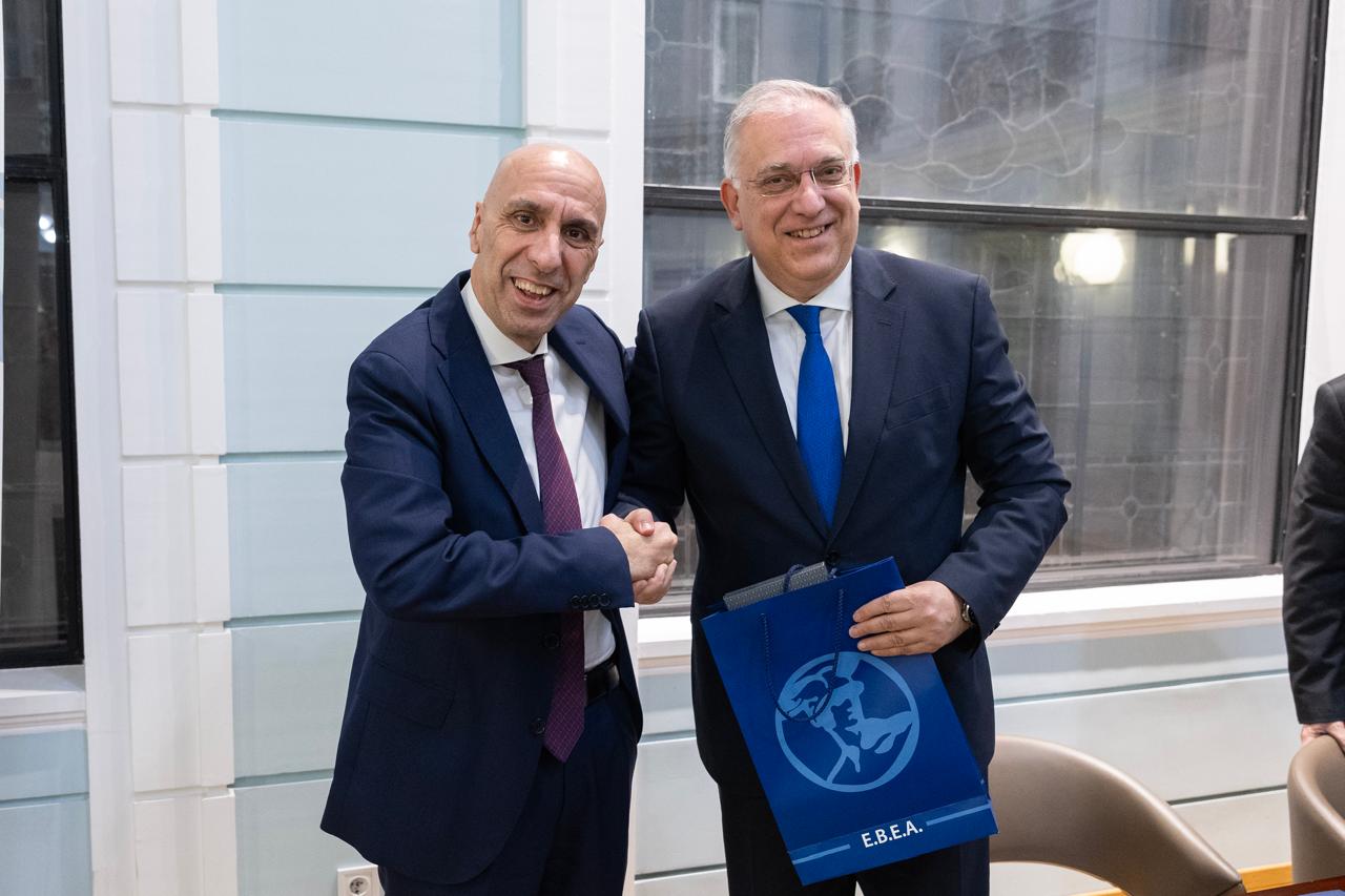 Two men are shaking hands, smiling, in a formal setting. One holds a blue bag with "E.B.E.A." printed on it.
