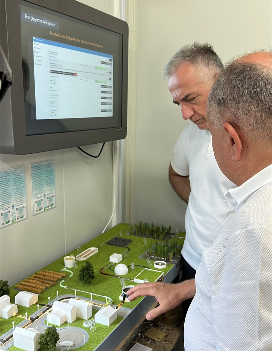 Two men are examining a model layout of a project, with a screen displaying data in the background.
